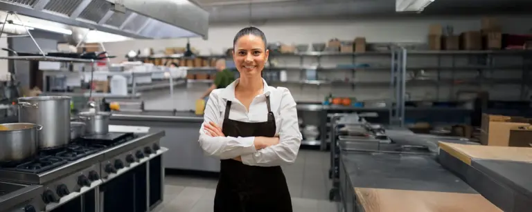 Köchin in professioneller Restaurantküche, lächelnd, mit verschränkten Armen, modernes Arbeitsumfeld, Edelstahlgeräte.