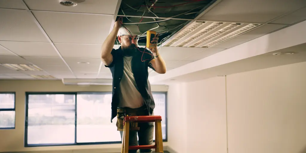 Elektriker auf Leiter prüft Kabel in abgehängter Decke eines Büros. Arbeitssicherheit, Installation, Technik.