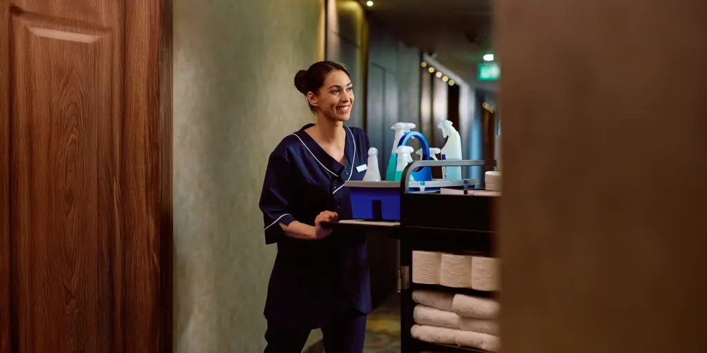 Putzkraft im Hotel mit Reinigungswagen, gefüllt mit Tüchern und Reinigungsmitteln, lächelnd auf einem Flur.
