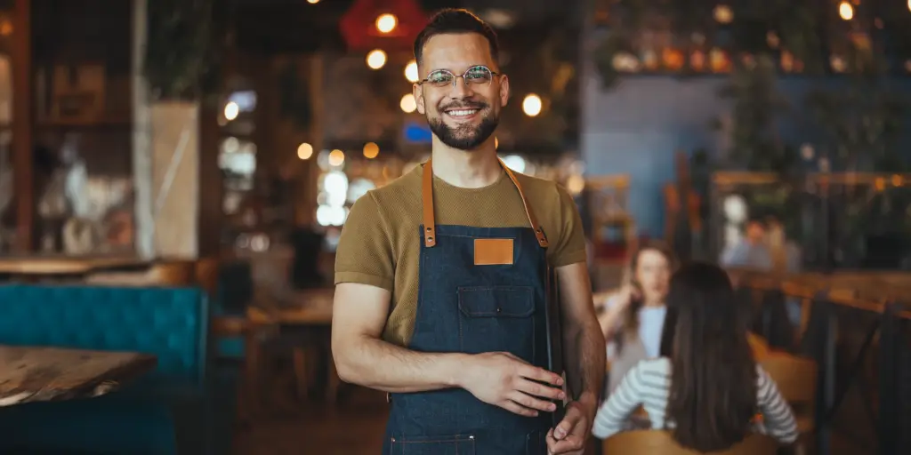 Ein lächelnder Kellner im Café trägt eine Schürze. Im Hintergrund sind Gäste an einem Tisch.